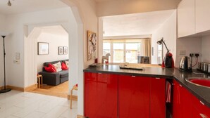 A modern kitchen with red cabinets, a black countertop, and a dining area with a grey sofa and red cushions. - 4guests-2br-privategarden-freeparking (Aviemore)