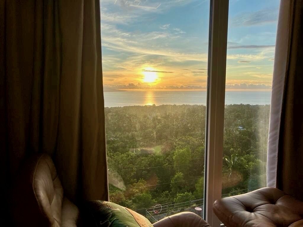A room with a large window offering a view of a sunset over a body of water and a forested area.