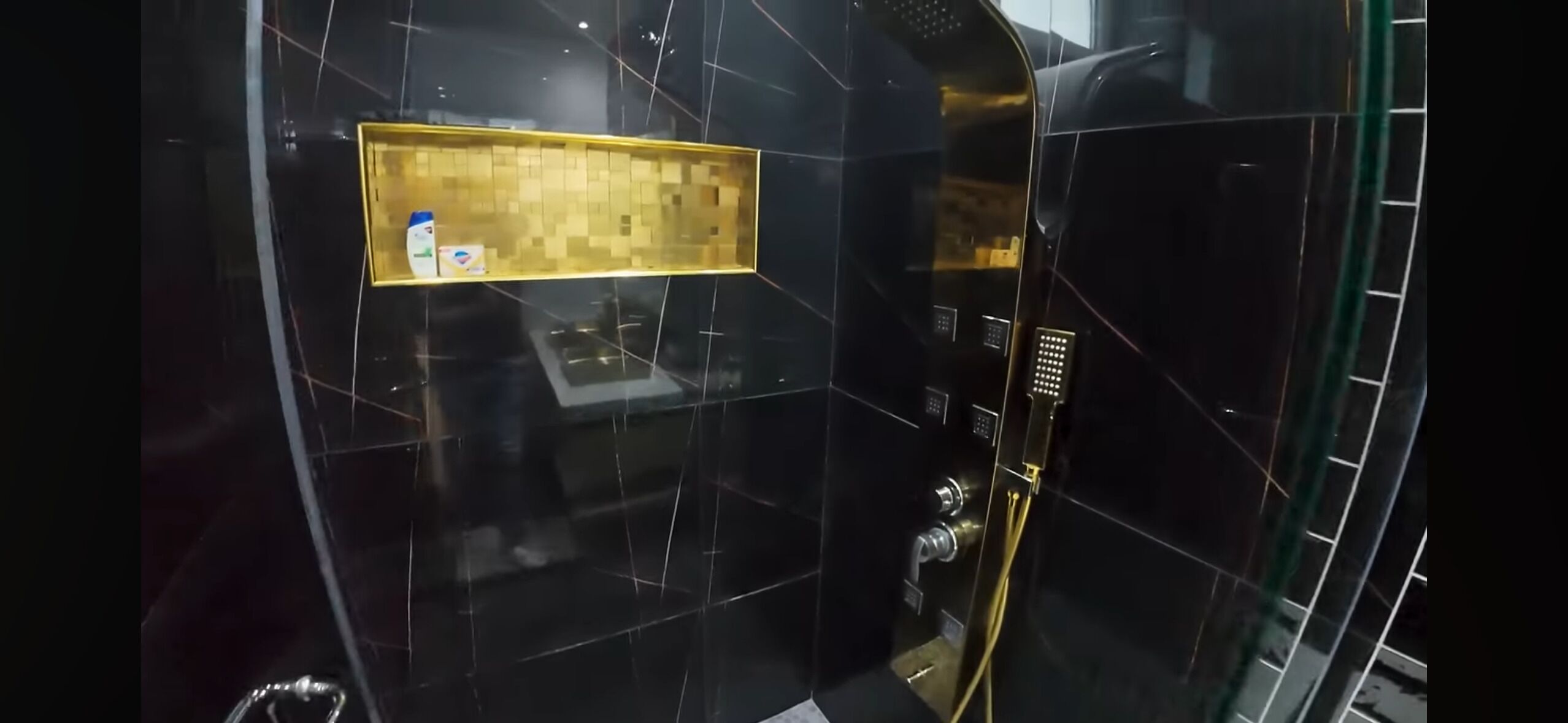 Um banheiro moderno com paredes revestidas de azulejos pretos, um espelho com moldura dourada e painel de comando do chuveiro.
