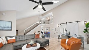 A spacious living room with a grey sectional sofa, a round coffee table, and a brown leather chair. A staircase leads to an upper level with a kitchen and dining area. - The Fairway Retreat at Vista del Verde (San Diego)