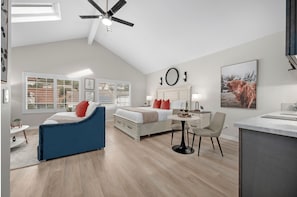 A modern bedroom with a bed, a desk, and a chair. There is a window with blinds, a ceiling fan, and a painting on the wall.