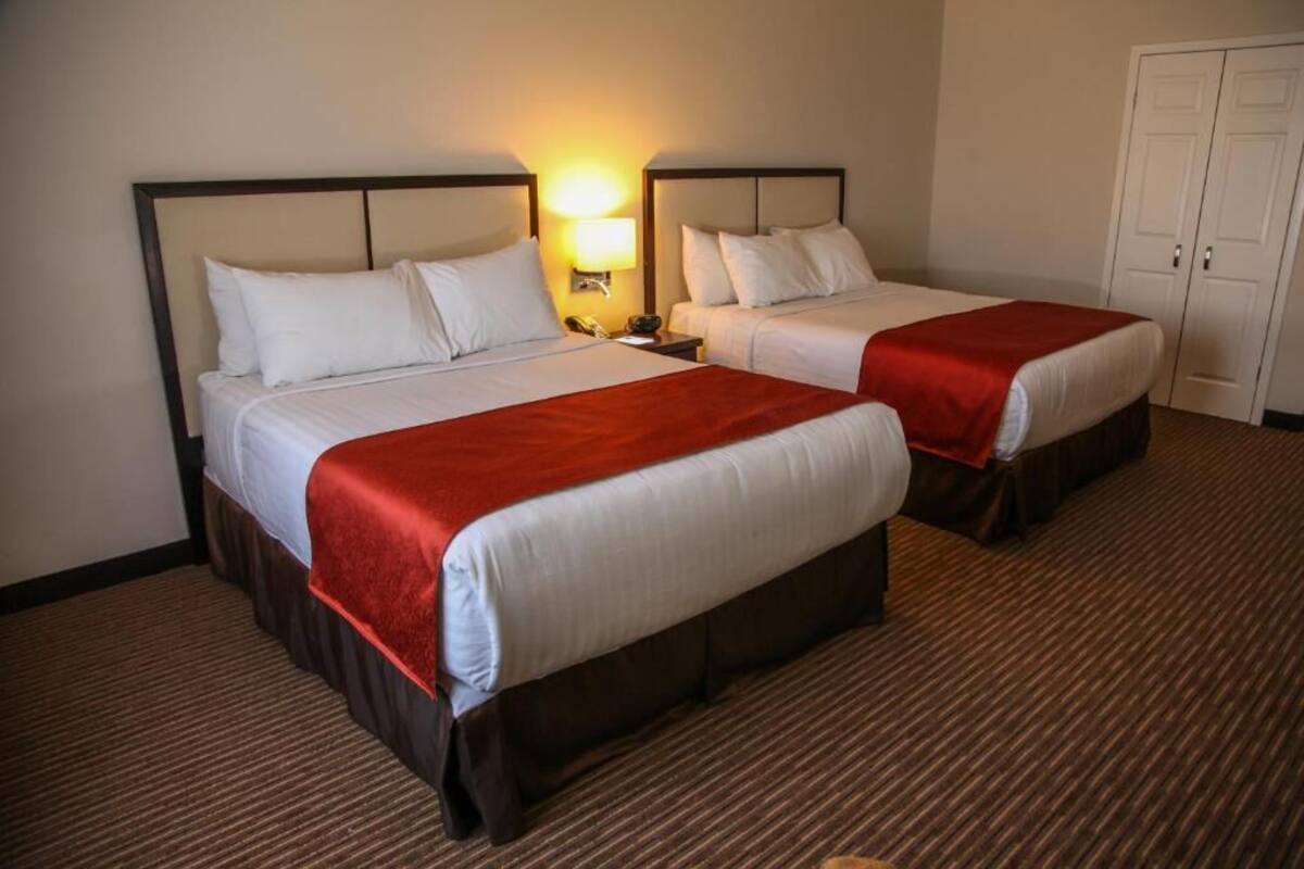 Ein Hotelzimmer mit zwei Betten, jeweils mit weißer Bettwäsche und roten Akzenten. Zwischen den Betten befindet sich ein Nachttisch mit einer Lampe, und in der Ecke ein Kleiderschrank.