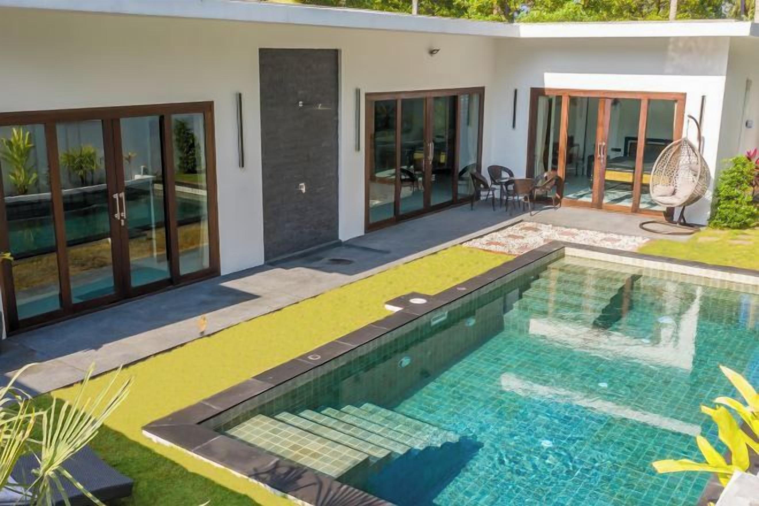 Una piscina exterior moderna con un elemento acuático de color azul transparente, rodeada por un patio con asientos y una entrada con puerta de cristal.