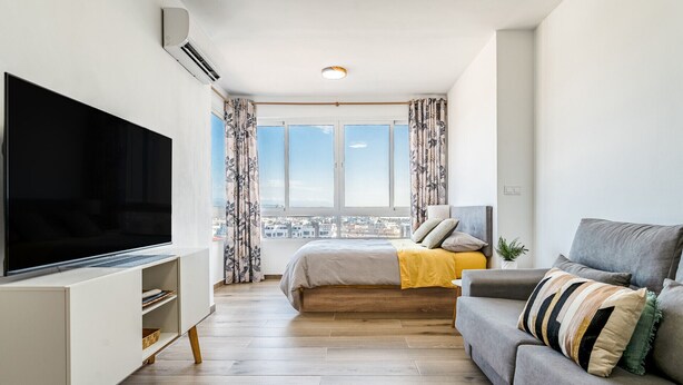 Un dormitorio moderno con un televisor grande, una cama con una manta amarilla y un sofá gris.