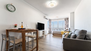 Una sala de estar moderna con un sofá gris, una mesa de comedor de madera con dos sillas, un televisor de pantalla plana y un amplio ventanal con vistas a la ciudad.