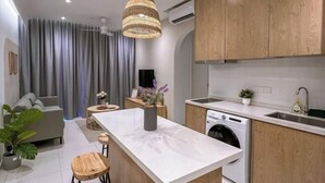 A modern kitchen with wooden cabinets, a white countertop, a built-in washing machine, and a dining area with a grey sofa and a small table. - Kenwingston Skyloft Apartment Doorsteps to Brt Subang USJ (Subang Jaya)