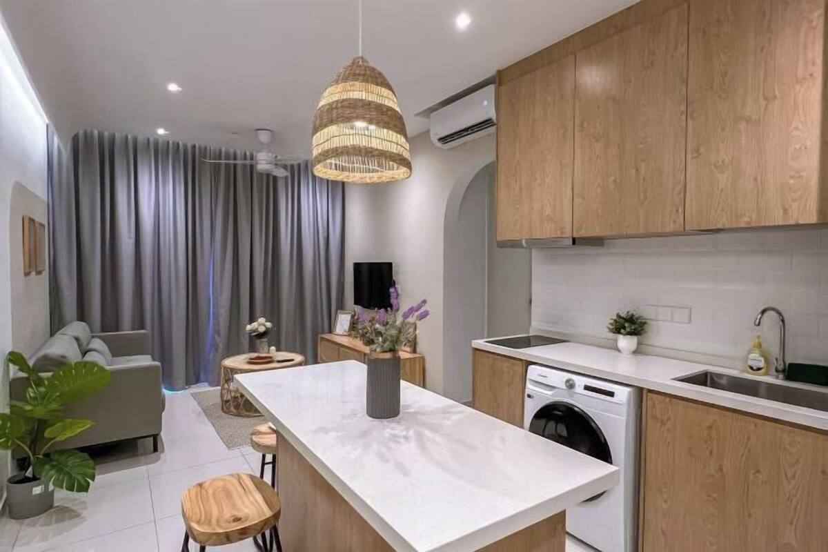 A modern kitchen with wooden cabinets, a white countertop, a built-in washing machine, and a dining area with a grey sofa and a small table.