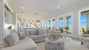 A spacious living room with a large sectional sofa, a patterned ottoman, and a view of the kitchen and balcony. - Featured in American Dream Home !  Stunning Oceanfront Views for Days !! (Galveston)