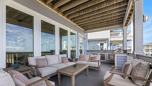 A covered outdoor seating area with sofas and a coffee table, wooden deck flooring, and large windows. - Featured in American Dream Home !  Stunning Oceanfront Views for Days !! (Galveston)
