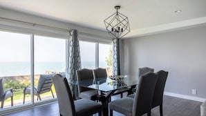 Un comedor con mesa y sillas de vidrio, ventanas amplias con vistas al océano e iluminación de techo geométrica.