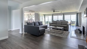 Un salón amplio con un sofá gris, un sofá color beige y una mesa de centro. Se observa una chimenea a la derecha y un ventanal grande con vista al océano.