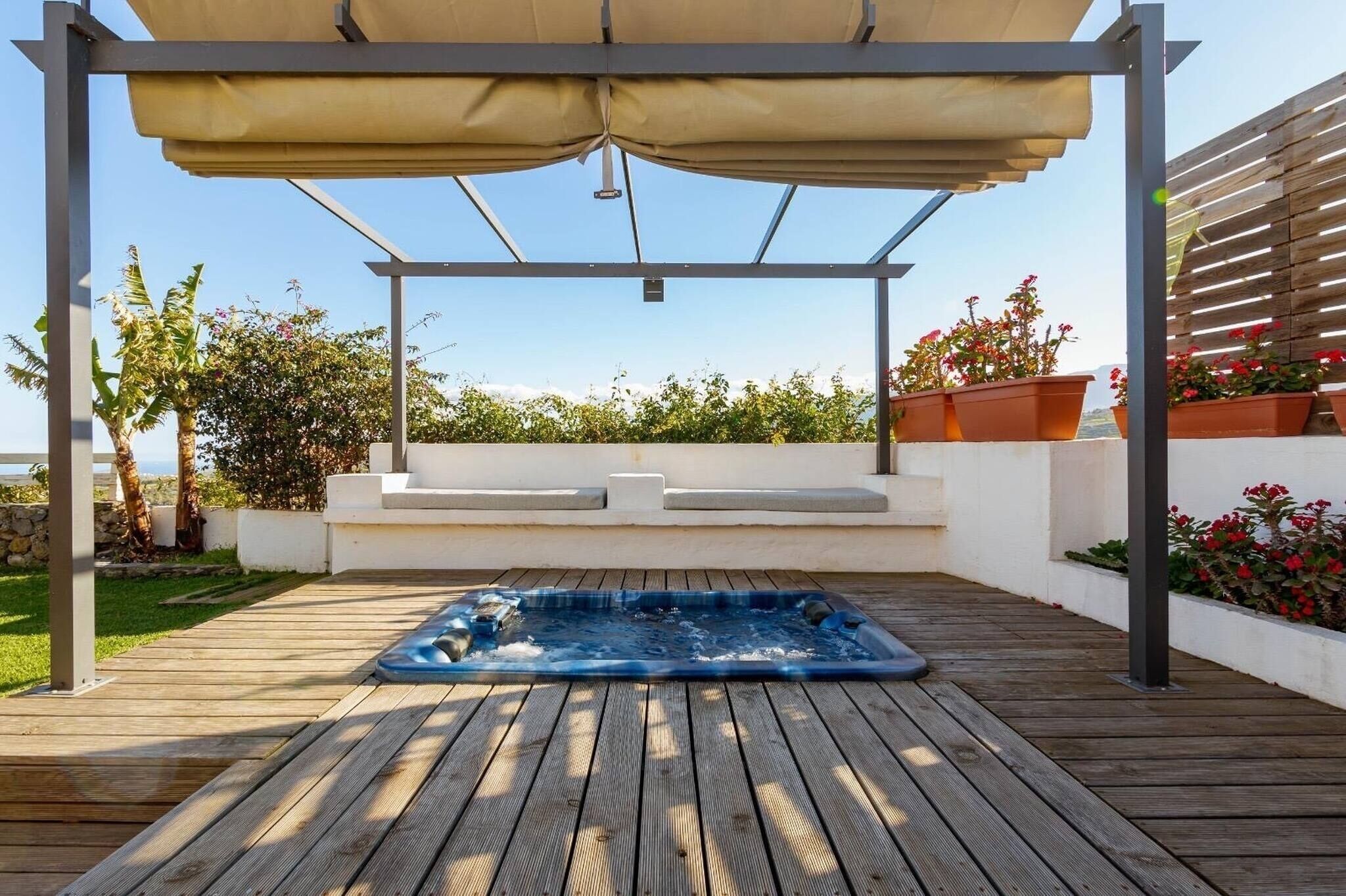 Una terraza de madera con un jacuzzi, una pérgola con toldo y plantas en macetas.