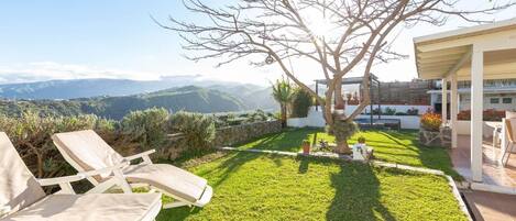 Un césped cuidado con un árbol, un patio con sillas y vistas a montañas distantes.