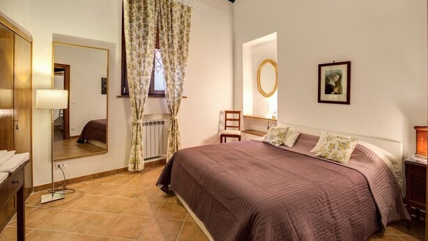 A bedroom with a large bed, a mirror, a window with curtains, a bedside table, and a wall-mounted lamp. - Lovely and cosy apartment nearby Navona Square (Roma)