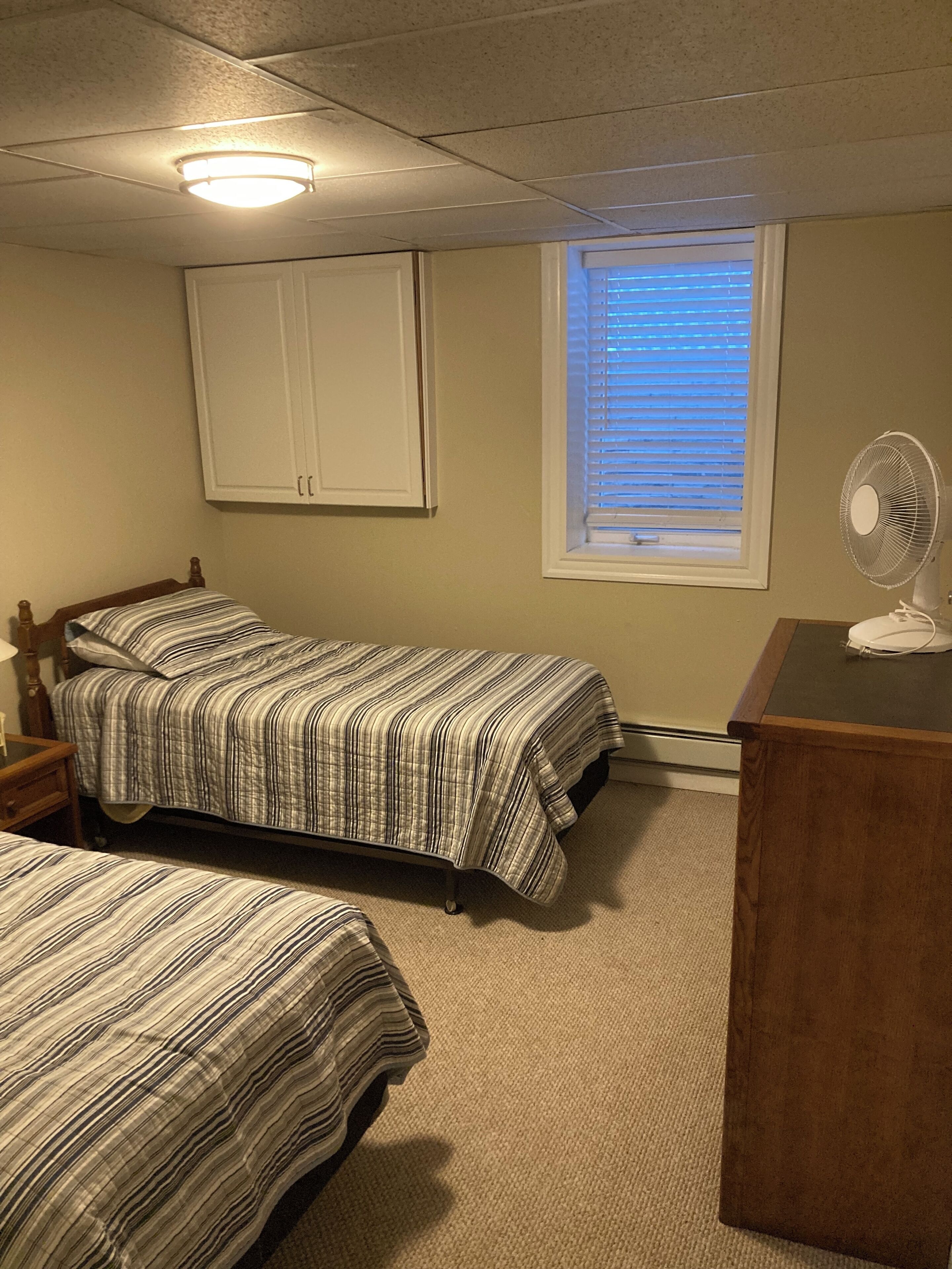 A bedroom with two beds, a window with blinds, a ceiling fan, and a wooden dresser.