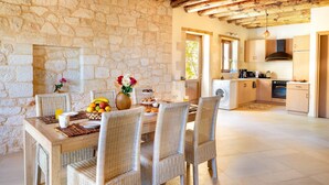 A rustic stone-walled dining area with wicker chairs, a wooden table, and a fruit bowl. A kitchen with modern appliances is visible in the background. - Villa Erontas Mini Pool and Heated Jacuzzi (Apokoronas)