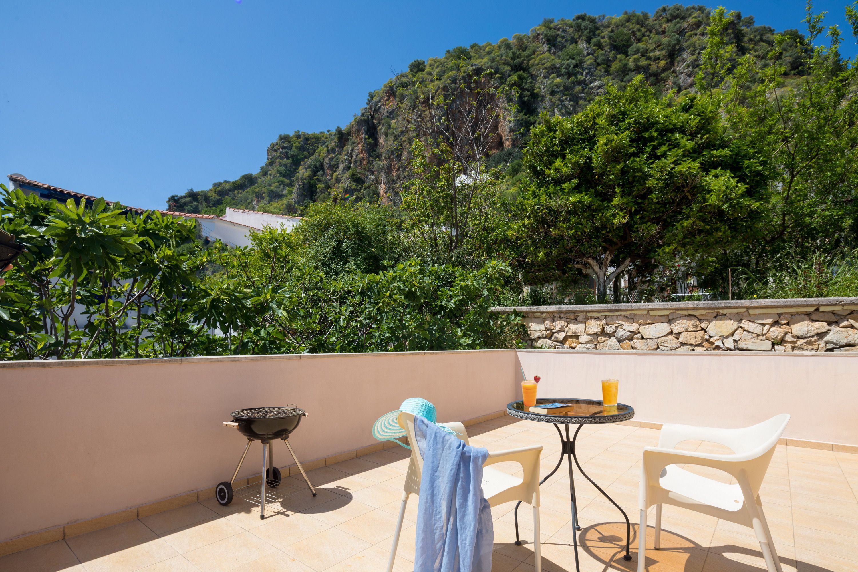 A terrace with a barbecue, two chairs, a table with a drink, and a view of a mountainous landscape.