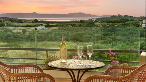 Un balcon avec des fauteuils en osier, une table avec une bouteille et deux verres à vin, et une vue sur un paysage côtier avec des montagnes et de la végétation.