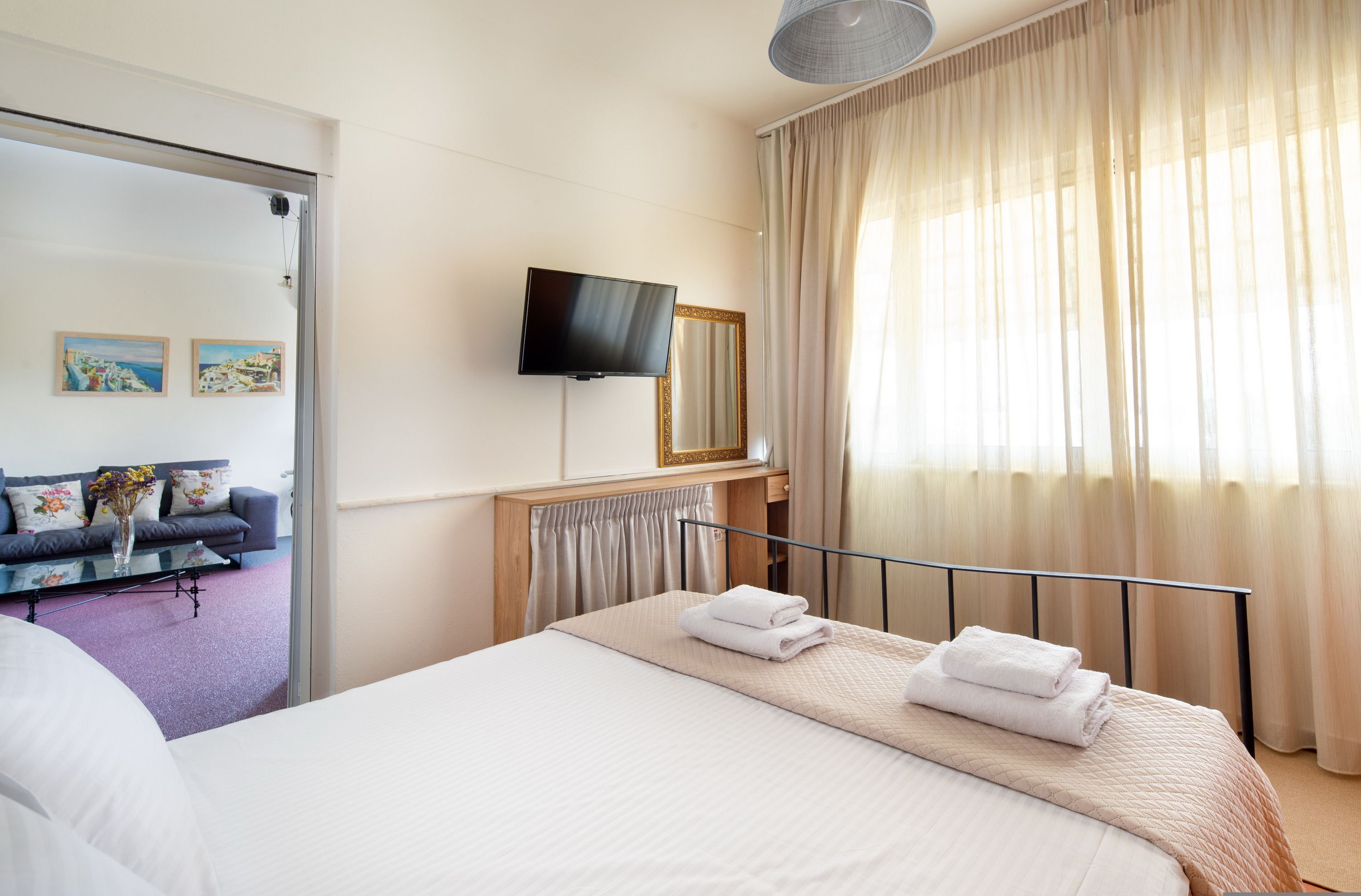 A hotel room with a bed, a TV mounted on the wall, a window with curtains, and a sofa with a floral vase.