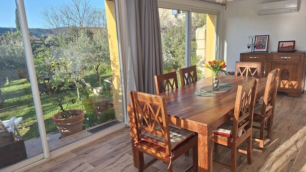A dining area with a wooden table and chairs, large windows with curtains, a ceiling fan, and an air conditioning unit. - House Rustrel between Ochres and lavenders 3 bedrooms (Rustrel)