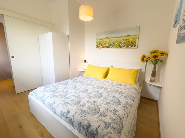 Un dormitorio con cama, mesita de noche, lámpara, un cuadro y un jarrón con flores.