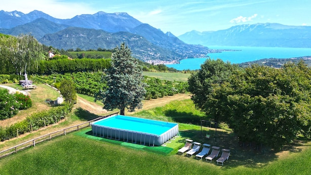 An aerial view of a property with a swimming pool surrounded by lush greenery, a wooden deck, and a view of a lake and mountains in the background. - Villa Anna Miralago in Puegnago sul Garda (Puegnago del Garda)