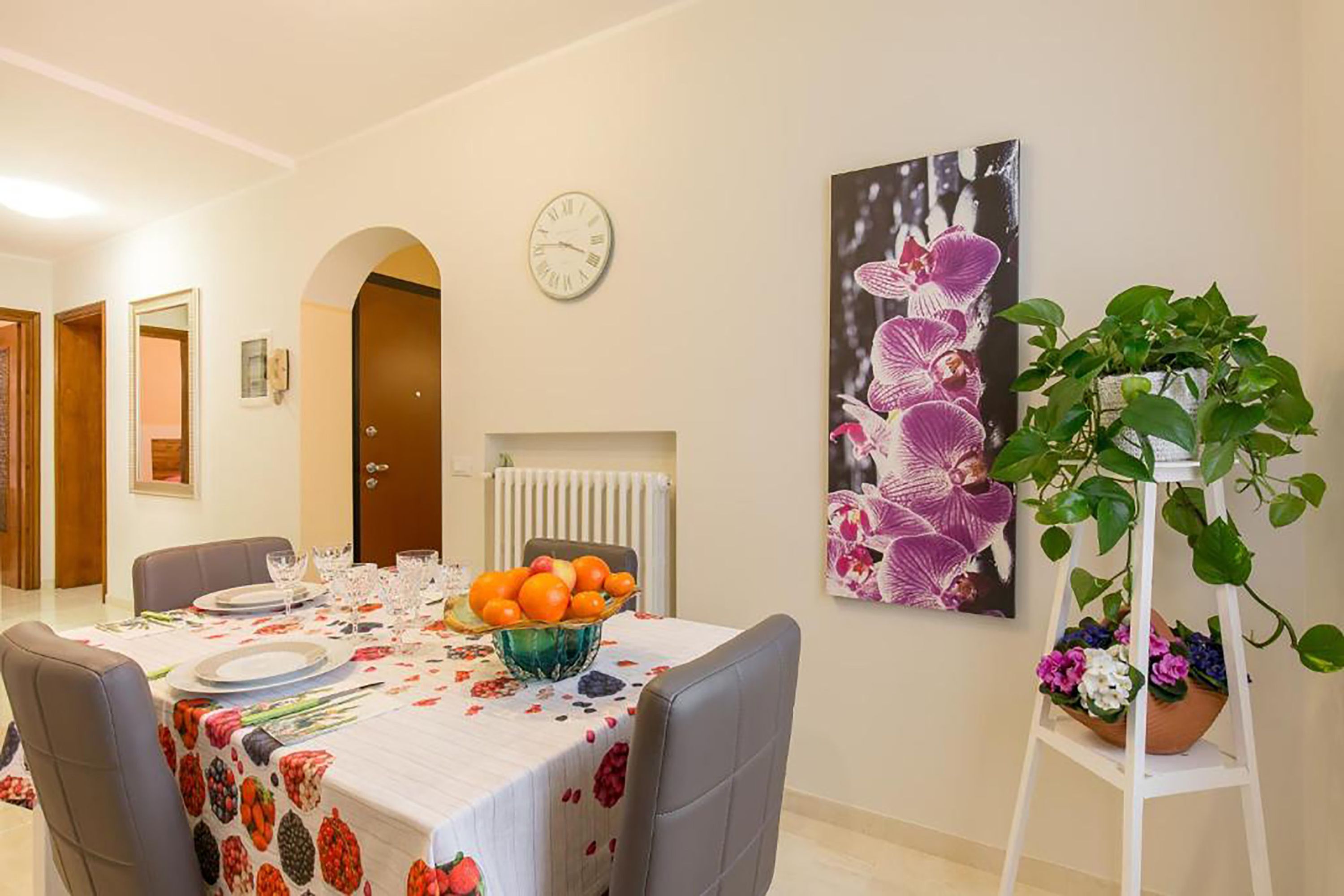 Un coin repas avec une table dressée pour quatre personnes, une horloge au mur et un grand tableau représentant une orchidée.