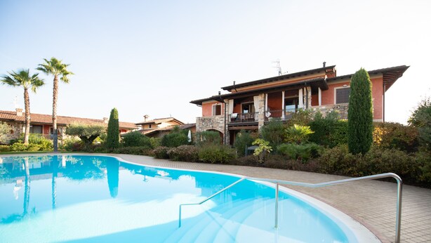 A swimming pool with a stone pathway leading to a house with a balcony. - Villaggio Tavaredo - App 02 (Polpenazze del Garda)