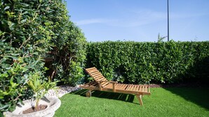 Una silla lounge de madera sobre césped artificial, rodeada por un seto cuidado y una planta en maceta.
