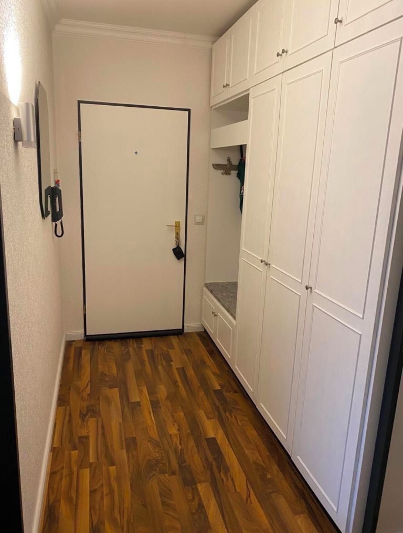 A hallway with wooden flooring, a white door, and white cabinets with multiple doors.