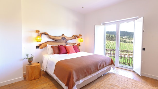 Un dormitorio con una cama grande, un cabecero de madera, un taburete de madera pequeño con una planta y vistas a un balcón con barandilla.