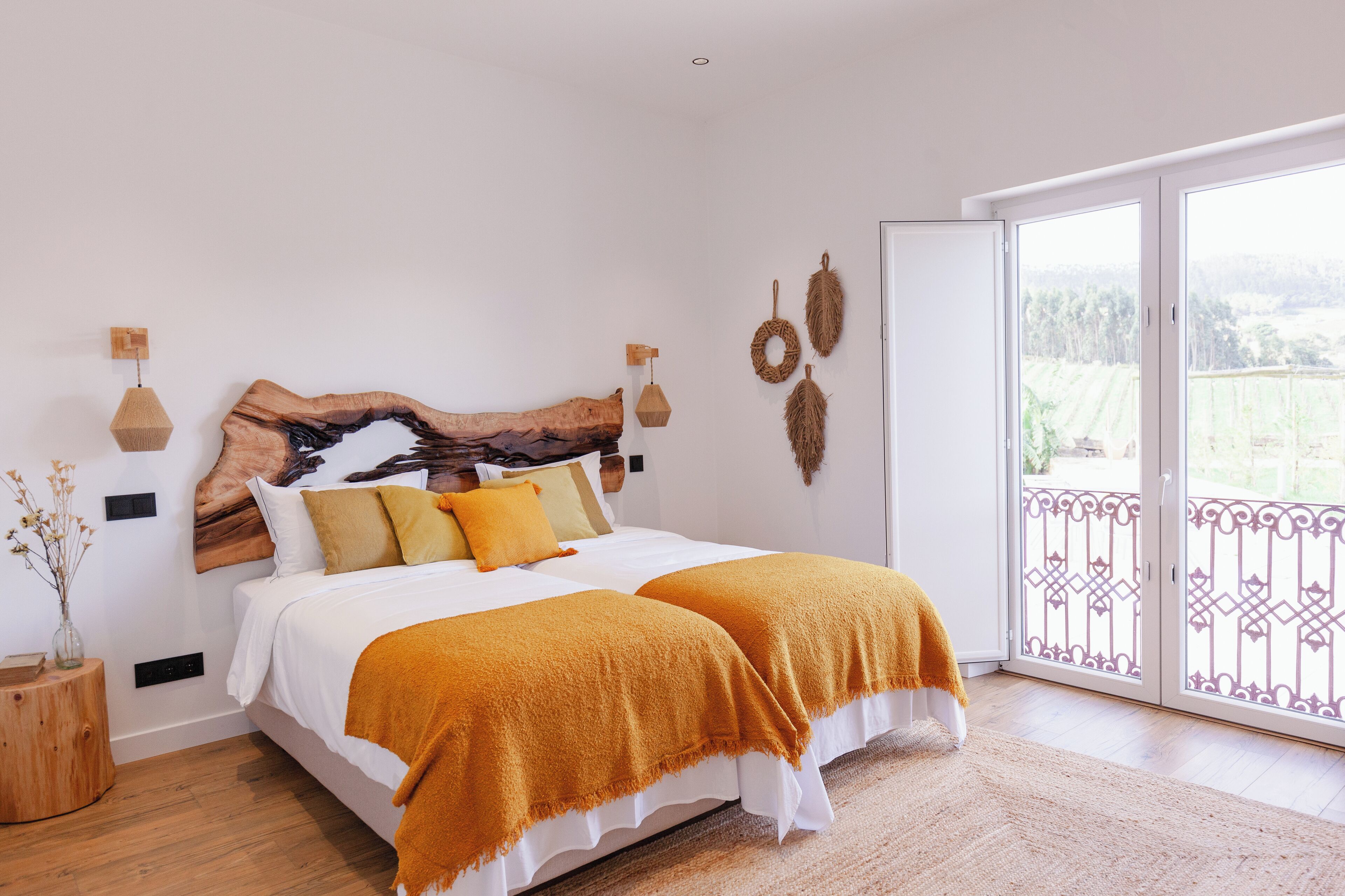 A bedroom with a wooden headboard, two beds with orange bedding, a wooden side table, and a large window with a view of greenery.