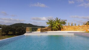 Una piscina con muro de piedra y palmeras, rodeada de césped y colinas al fondo.