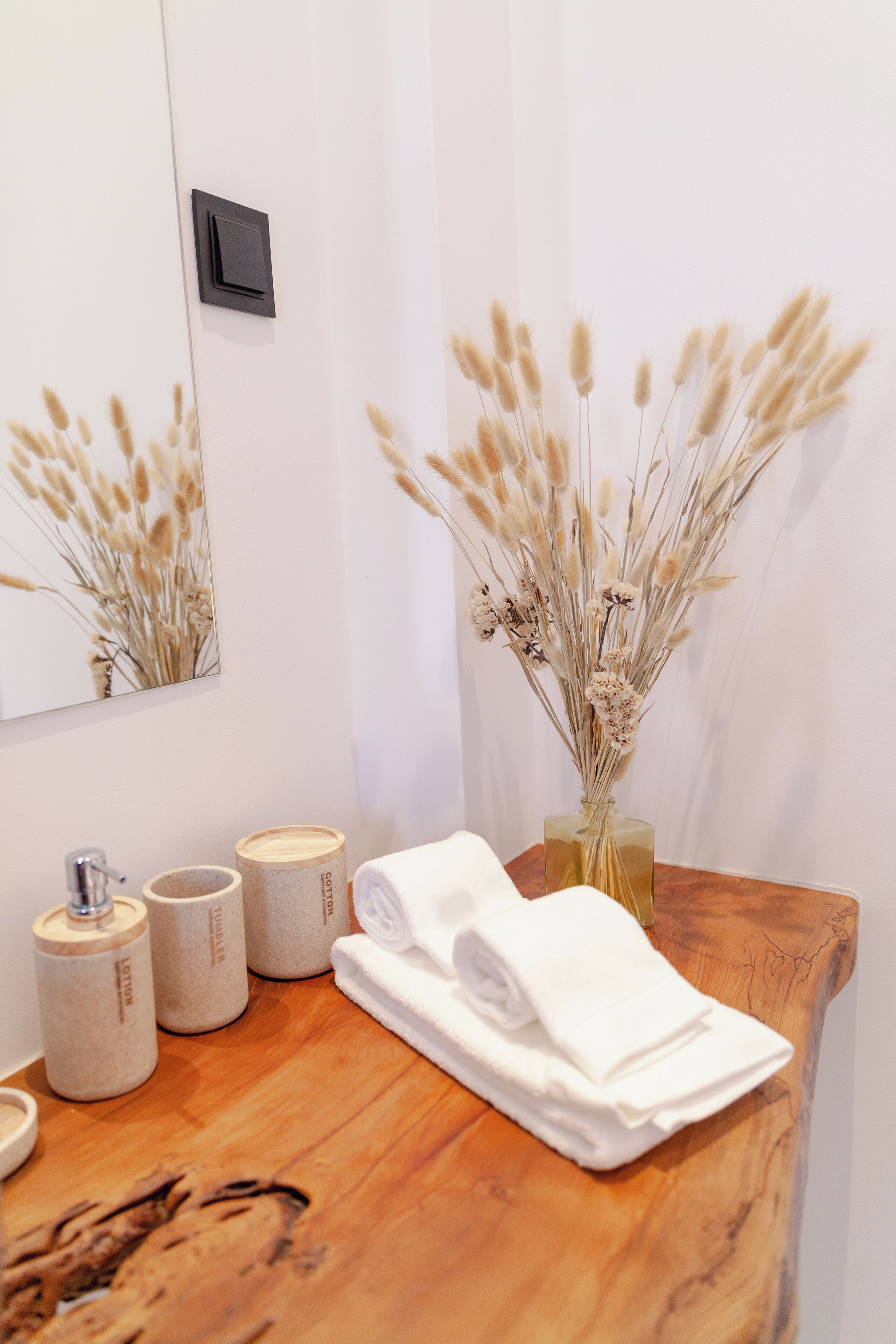 A bathroom with a wooden vanity, a mirror, a vase with dried flowers, and toiletries.