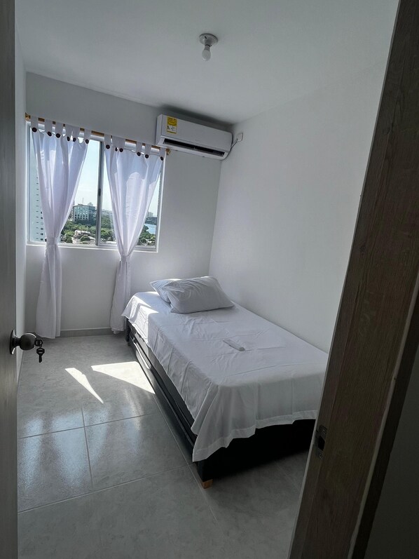 Een kleine, schone kamer met een bed, een raam met gordijnen en een airconditioning.