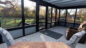 A covered patio with a wooden table and cushioned seating, large windows offering a view of a yard with trees and a fence. - Cozy cabin on the Creek with WiFi and AC in delightful Belton (Belton)