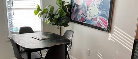 A dining area with a table and chairs, a potted plant, a painting on the wall, and a patterned rug.