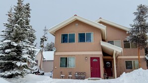 A two-story building with a red door, a covered porch, and a snow-covered landscape. - 3/3 Townhome in Fraser just steps from the Black line! (Granby)