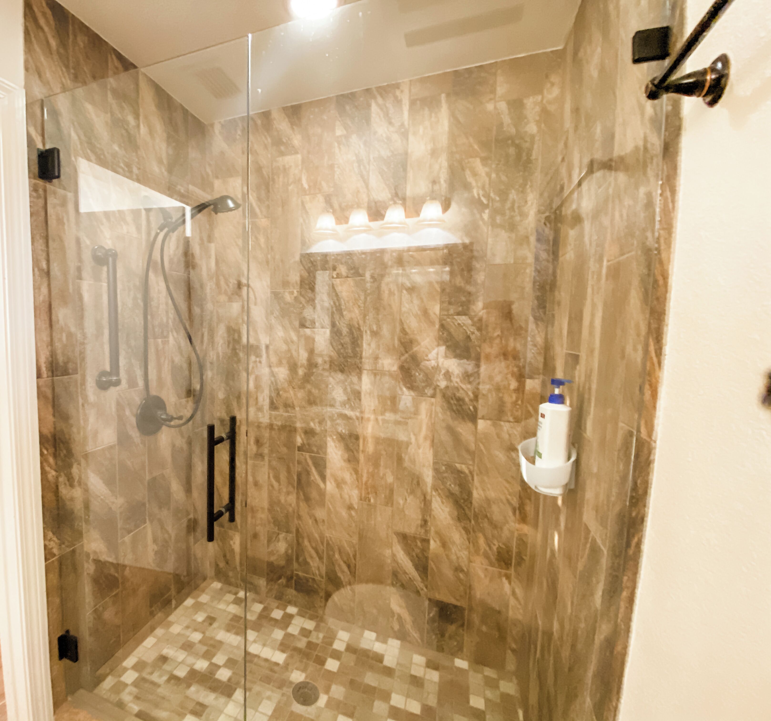 Une douche fermée par des parois de verre, avec un carrelage à motifs géométriques, une douchette à main et un luminaire fixé au mur.