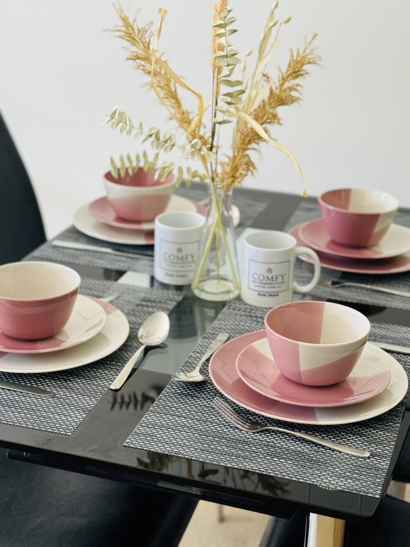 A table set with pink and white bowls, spoons, and a vase with dried plants.