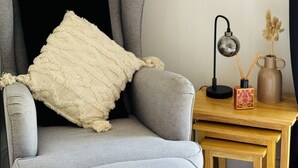 Un sillón gris con una manta blanca y un reposapiés a juego. Una mesita de madera con una lámpara y un jarrón de flores secas está al lado de la silla.