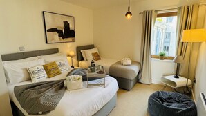 Habitación de hotel con una cama grande, otra más pequeña, un sillón de saco, una mesita de noche con lámpara, una ventana con cortinas y un cuadro en la pared.