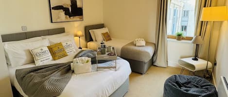 A hotel room with a large bed, a smaller bed, a bean bag, a bedside table with a lamp, a window with curtains, and a painting on the wall.