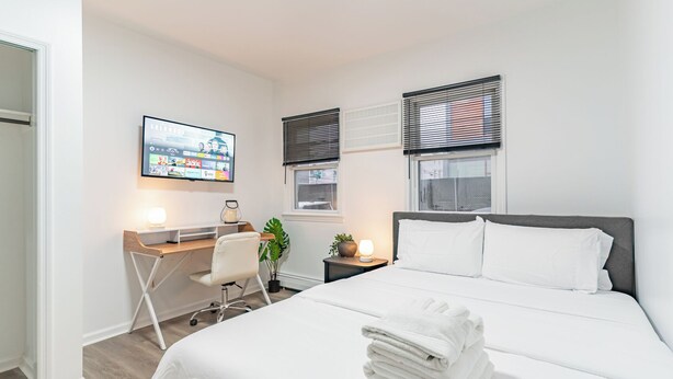 A hotel room with a bed, a desk, a chair, a TV, and a window with blinds. - Transit Oasis: EWR 8 Min/Penn with Free Parking (Newark)