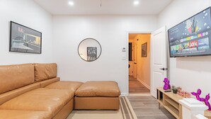 A modern living room with a brown leather sofa, a round wall clock, a flat-screen TV mounted on the wall, and a wooden bench with decorative items. - Transit Oasis: EWR 8 Min/Penn with Free Parking (Newark)