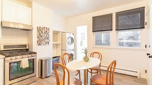 A compact kitchen with a stove, oven, and washing machine, a dining table with chairs, and a window with blinds. - Transit Oasis: EWR 8 Min/Penn with Free Parking (Newark)