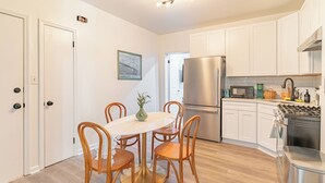 A modern kitchen with white cabinets, a stainless steel refrigerator, a gas stove, and a dining area with a round table and wooden chairs. - Transit Oasis: EWR 8 Min/Penn with Free Parking (Newark)