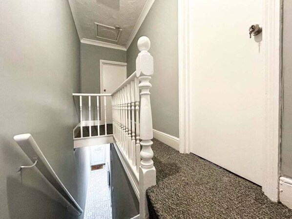 A narrow hallway with a white door at the end, a white railing with a baluster, and a carpeted floor.
