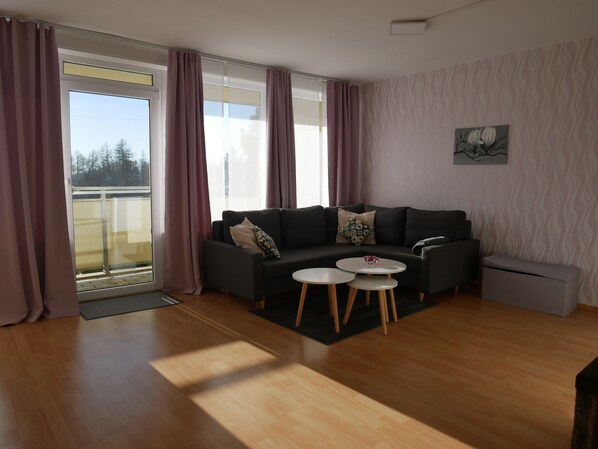 A modern living room with a dark grey sofa, two white side tables, and a view of a balcony through large windows with pink curtains. - Panoramic Hohegeiß, Apartment 374 with swimming pool and sauna (Braunlage)