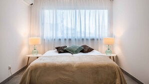 Hotellihuoneessa on sänky, kaksi yöpöytälamppua, ikkuna, jossa on läpikuultavat verhot, ja seinälle asennettu ilmastointilaite.